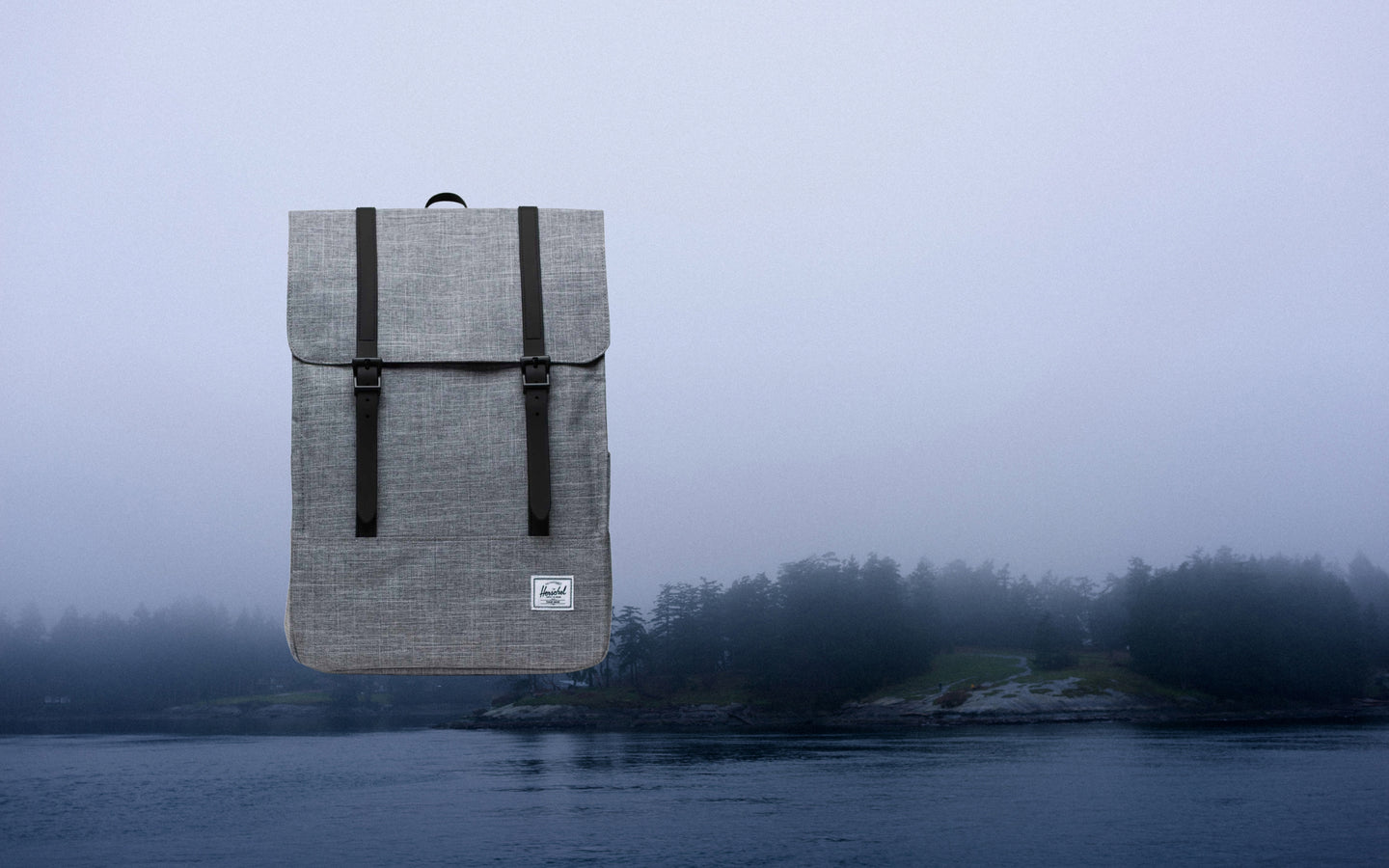 Herschel Survey Backpack in Raven Crosshatch. A gray heathered backpack with two black rubber straps on the front flap.